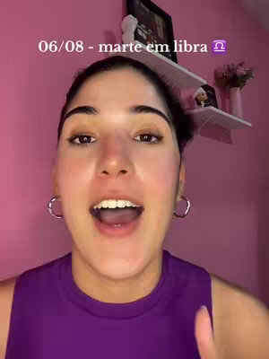 A woman with dark hair and hoop earrings is in the foreground, emoting and speaking. She wears a purple top. Behind her is a pink wall with a white shelf, adorned with a small framed picture and a vase of flowers.