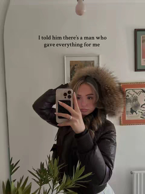 A woman taking a selfie in front of her mirror. She is wearing a brown winter coat with a furry hood and has her phone raised covering most of her face.  Text overlay: 'I told him there’s a man who gave everything for me'.