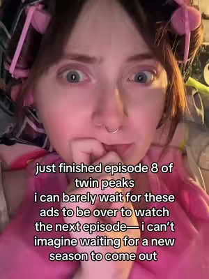 A close-up shot of a person with light skin, looking directly at the camera with wide, excited eyes. They have dark hair with bangs and are wearing pink hair rollers. Their index finger is touching their lips, as if they are deep in thought or slightly worried, and their cheeks are flushed pink. The background is blurred, suggesting a close-up shot with soft lighting. The person is wearing a light pink top.