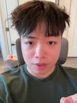 The video cover features a medium shot of an Asian male with messy dark hair and a neutral expression. He is seated, likely indoors, wearing a simple green shirt. The background includes hints of a room with white walls and visible items like a chair and a part of a pink stuffed animal.