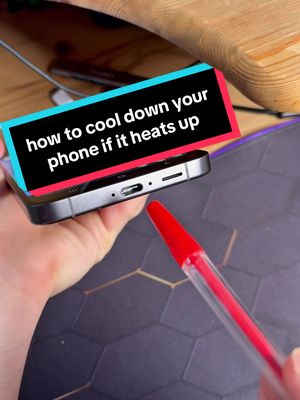 A hand holding a smartphone, with a red and clear pen pointing towards the bottom of the phone. The phone is black, and it has a text overlay that reads 'how to cool down your phone if it heats up.' The background showcases a wooden surface, a black surface with lines, and part of a computer mouse.