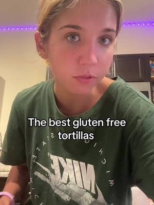 A woman is centered in a close-up shot, looking directly at the camera. She has blonde hair and is wearing a green t-shirt with a Nike logo on it. A black text box is displayed on the bottom of the screen with white text that says "The best gluten free tortillas". The background is slightly blurred, with purple lights in the top right corner.