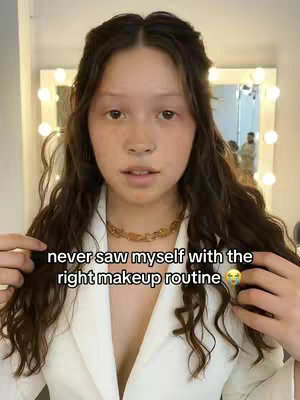 A young woman with long, wavy brown hair and a gold chain necklace poses in front of a bright makeup mirror. She wears a white blazer, and the overlay text is centered on her face.