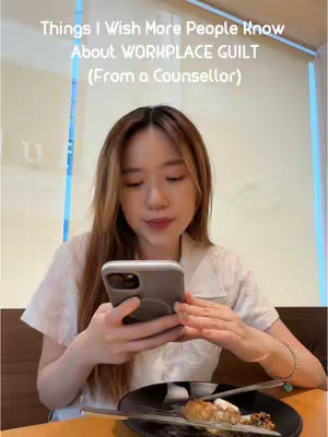 A young woman with long, light-brown hair is seated indoors, looking down at her phone. The background includes a window, and a table is partially visible in front of her. She is holding her phone in her hands, and there is a plate of food behind her, suggesting a relaxed, casual setting.