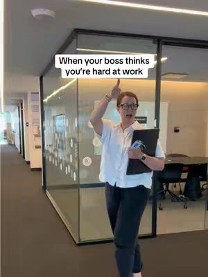A woman in business attire is standing in an office setting, raising her hand and pointing upwards. She is wearing glasses, a white shirt, dark pants, and holding a black folder with a phone tucked in. The background shows glass walls and office corridors with doors.