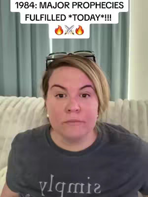 A woman with light skin and short, highlighted blonde hair appears in a selfie-style video. She is wearing a dark gray t-shirt with text. Above her head, text overlays read "1984: MAJOR PROPHECIES FULFILLED *TODAY*!!!" with flame and sword emojis. She is sitting on a cream-colored couch in front of light-blue curtains.