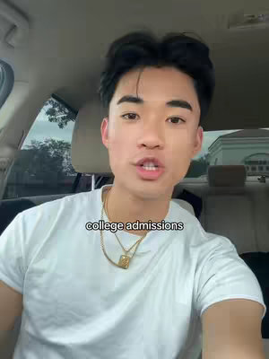 A young Asian man is seated inside a car, gazing directly at the camera. He is wearing a white t-shirt and a gold chain. The background shows the interior of the car, with headrests visible. Text overlayed reads 'college admissions'. The lighting is natural, and the composition is a medium shot, focusing on the man's face.