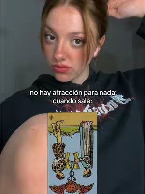 A person with brown hair in a black hoodie is holding their hair up and looking at the camera with a slightly pursed expression. A tarot card is in view, upside down, with text overlayed.