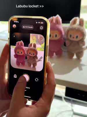 A person is holding a smartphone showing a photo of two Labubu dolls in a soft pink and brown/tan color holding a small white flag, and the case is gold in color. Behind the phone, a blurry background with three Labubu dolls in a row. The foreground consists of the hand holding the phone.