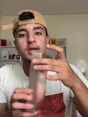 A man in a white t-shirt and a tan baseball cap is holding a clear bottle in front of him. The background includes shelves with perfumes and colognes.