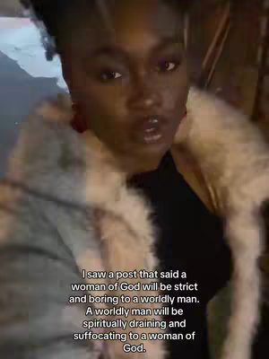 A woman with dark skin is centered in the frame, facing the camera. She has soft features, with a slightly open mouth. She's wearing what appears to be a black top and a light-colored, fuzzy coat, giving a soft texture around her shoulders. The background is dimly lit, adding to the sense of depth and enhancing the subject. Text overlay on the bottom reads.