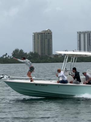 A man in a light-colored shirt stands on the front of a motorboat with his arm outstretched. Several other people are on the boat, and buildings are in the background. The water is light azure, the sky overcast.