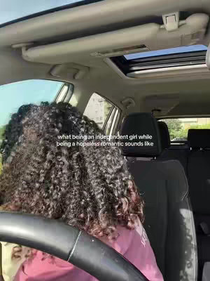 The image shows a person with curly hair, seen from the back, sitting in the driver's seat of a car. The person is wearing a pink top and holding the steering wheel. The car interior includes a sunroof, a dashboard, and the headrest of the front passenger seat. Text overlay reads 'what being an independent girl while being a hopeless romantic sounds like'.