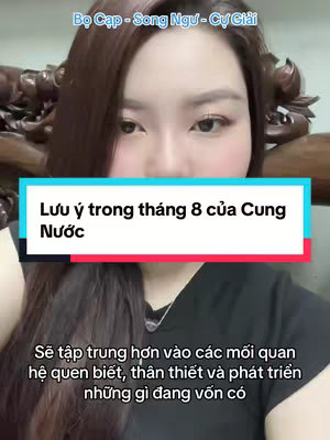 A woman with long, dark hair is centered in the frame, with a brightly colored sign overlay that reads, 'Lưu ý trong tháng 8 của Cung Nước'. The background is slightly blurred, featuring what appears to be wooden furniture. The woman is facing the camera.