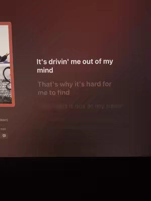 The image is a screenshot of a black screen with white lyrics from a song. The lyrics 'It's drivin' me out of my mind' are written on the top half of the screen, followed by 'That's why it's hard for me to find' beneath it. The bottom right corner displays more text that is illegible. The background suggests a dark, simple setting that is in focus.
