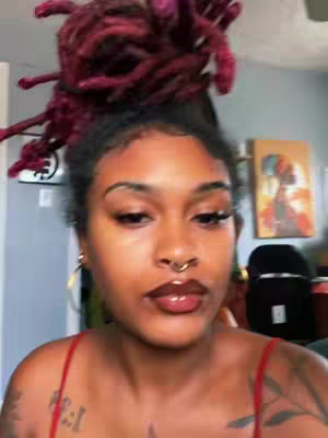 A close-up shot of a woman with dark skin, styled reddish-burgundy dreadlocks piled high on her head, and a septum ring, is looking into the camera. The lighting is natural and the background is blurred, showing elements of a room.