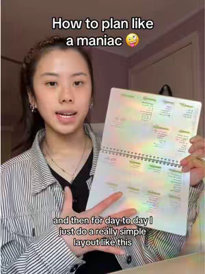 A young woman with dark hair is holding a planner open and displaying its contents while looking directly at the camera. She wears a striped button-up shirt. The background is out of focus, but suggests an interior space.