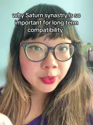 A selfie-style video cover image showcasing a woman with dark hair and glasses looking directly at the camera. Text overlayed at the top states, 'why Saturn synastry is so important for long term compatibility'. The woman has lipstick on and is likely inside..