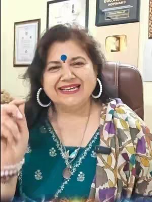 A woman with dark hair, wearing large hoop earrings, a green top, and a patterned scarf. She has a bindi on her forehead and a necklace. She is smiling and gesturing with her right hand. The background includes framed certificates and a gold YouTube play button.