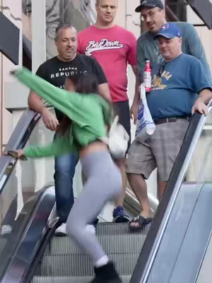 A woman appears to be falling on an escalator, in front of a group of men who appear to be recording.