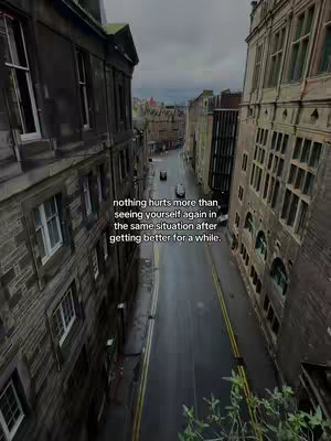 An aerial shot of a narrow street flanked by tall, aged buildings. The street is a dark asphalt, with yellow painted lines dividing the lanes. The buildings are dark, made of stone or brick, with numerous windows. The sky is overcast, contributing to a muted color palette of grays and browns. Text overlay provides a message about feeling stuck in a negative situation.
