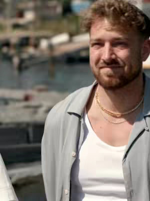 The cover image shows a man with light skin, short light brown hair and a beard. He is wearing a light gray button-down shirt over a white undershirt, and a gold necklace. He is smiling and facing slightly to the side, with a background of blurred buildings and a body of water.