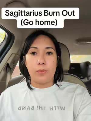 A woman is in a car, looking straight at the camera, with the added text overlayed on the top. The text reads"Sagittarius Burn Out (Go home)". The woman is wearing a white top, her hair is black. It is daytime, so the car is lit by natural sunlight.