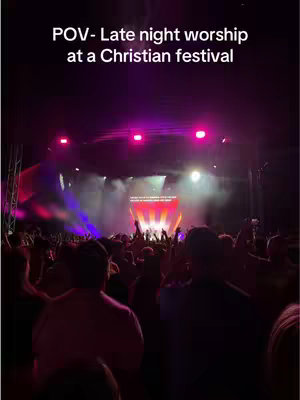 The image shows a live music event at a nighttime church festival. The point of view (POV) is from within the crowd, looking toward a brightly lit stage with a large screen. The lighting includes vibrant purples and pinks. The foreground is filled with the backs of people's heads and raised hands as they participate in the event. A text overlay at the top reads 'POV - Late night worship at a Christian festival'.