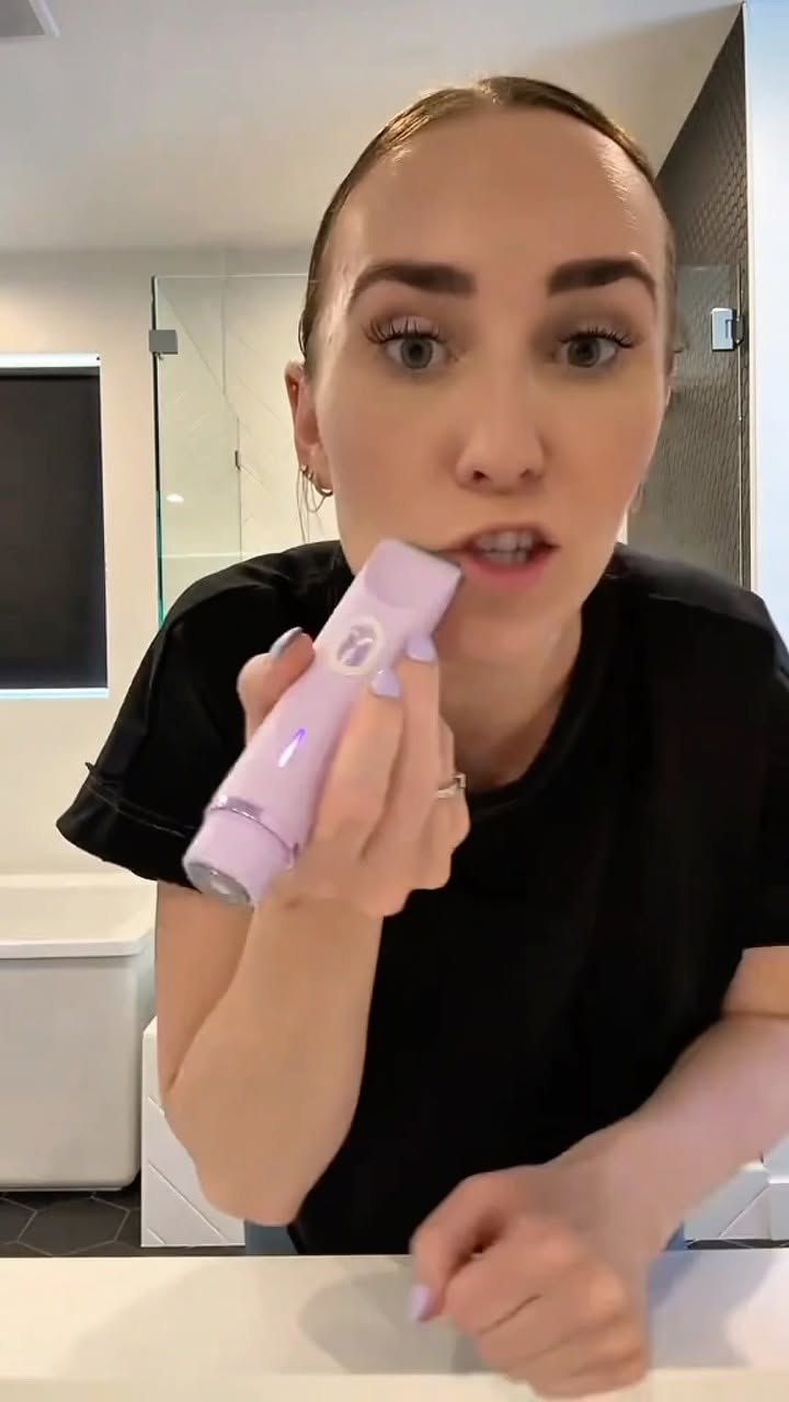 A woman is shown close-up in a bathroom setting, holding a lavender-colored hair trimming tool. The composition focuses on her face and hands as she demonstrates the use of the device.