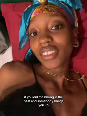 A medium shot features a woman lying down, likely in bed, looking towards the camera. She's wearing a black top and a scarf/head covering with a colorful floral pattern and blue accents. The background shows a red pillow. Text overlayed at the bottom reads, 'if you did me wrong in the past and somebody brings you up'.
