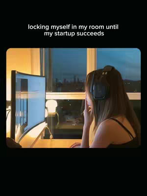 A woman is seated at a desk in her room, illuminated by a monitor and a desk lamp. She's wearing headphones. The room has a window with buildings visible in the distance. The text 'locking myself in my room until my startup succeeds' is overlaid at the top.