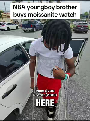 A man with dreadlocks and a white t-shirt is standing outside next to a car. He is holding a crutch and wearing a brace on one leg. Text overlays include "NBA youngboy brother buys moissanite watch", and details about profit and an instruction to click "HERE."