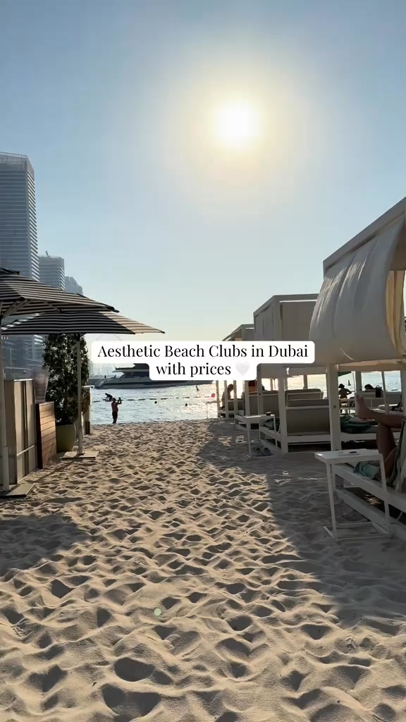 A shot of a sandy beach on a sunny day, with cabanas to the right and tall modern buildings in the background. The sun is directly overhead, creating a bright, high-contrast scene.