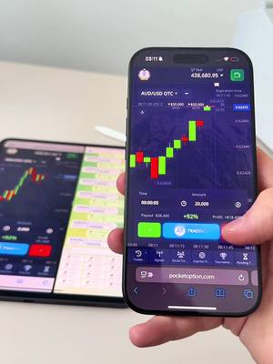 A person holds a smartphone displaying a trading app, with a tablet showing a similar interface in the background. The smartphone screen shows a candlestick chart with green and red bars, indicating market activity. The background is a light-colored surface, likely a desk.