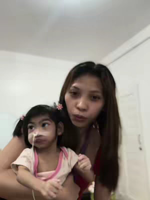 A woman holds a young child in her arms. The child has a nasal cannula. The woman is looking directly at the camera, and the background is a plain room.