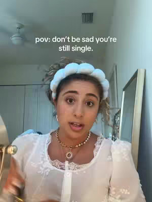 A woman with curly hair and a light blue headband is centered in the shot, looking directly at the camera. The background includes a ceiling fan, a light-colored wall, and a mirror. The woman is wearing light makeup, a white lace top, and a necklace with a sun pendant. The text overlay in the image reads, 'pov: don't be sad you're still single.'