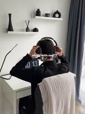 A person wearing headphones is shown from behind, seated at a desk. The room is minimalist, with white walls and shelves, adorned with black decorative items. Text overlayed on the image reads "study with me."