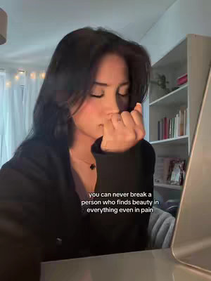 A young woman with dark hair and a black top is shown in a softly lit interior. She's holding her hand up towards her face, eyes closed, in a contemplative pose. The background includes a white bookshelf and a neutral-colored wall and ceiling. Text overlaying the image includes a quote about finding beauty in everything.