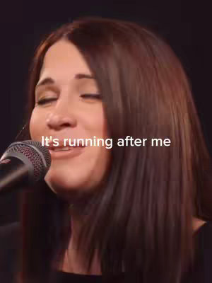 A woman with long brown hair is singing into a microphone with her eyes closed. Text overlayed on the cover image reads, "It's running after me."