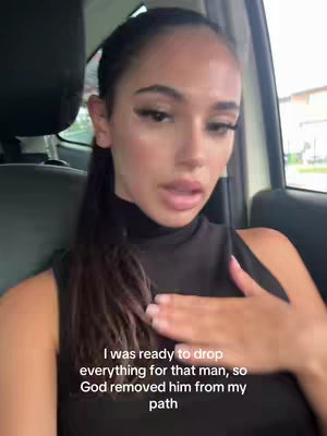 A woman with long dark hair and dark makeup is seated inside a car. She is wearing a dark, form-fitting sleeveless top. Her expression is serious, with her right hand touching her chest, and her mouth is slightly ajar as if she is speaking. The background shows parts of the car interior, with a blurred view of an outdoor scene visible through the car window.