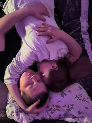 A young couple lies on a bed oriented with one person's head at the bottom and the other at the top of the image, with one playfully leaning in. The woman on top seems to be kissing the man on the other, both resting on a floral and patterned fabric. The composition is intimate and soft, using natural and ambient lighting.