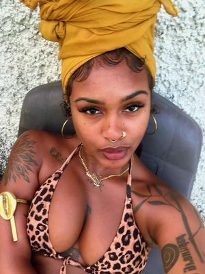 A selfie-style shot of a woman with dark skin, wearing a leopard print bikini top, gold jewelry, including hoop earrings, a nose ring, and a necklace. Her hair is covered by a yellow turban. She has visible tattoos on her arm and shoulder. The background appears to be an out-of-focus outdoor setting.