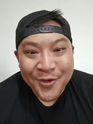 A close-up shot of a man's face, showing him with a slight smile and open mouth, as if speaking. He wears a black baseball cap and a black shirt, set against a plain, light-colored background. The lighting is even, highlighting the man's features.
