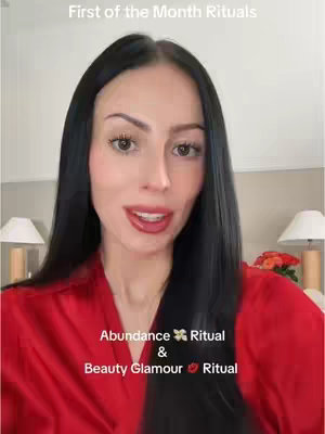 A woman with long dark hair is centered in a selfie-style video featuring the top half of her body. She is wearing a red robe, and two bedside lamps are behind her. Text overlays read \"First of the Month Rituals\" above, and \"Abundance Ritual & Beauty Glamour Ritual\" below.