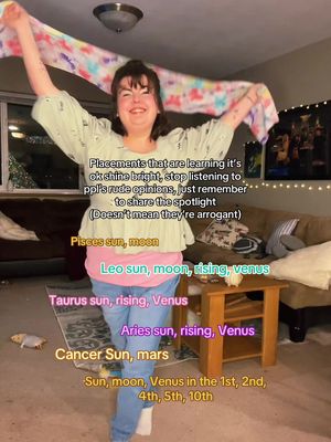 A woman stands with arms raised, holding a colorful scarf, in front of a living room setting. Text overlays list astrological placements, and positive message regarding self-expression.