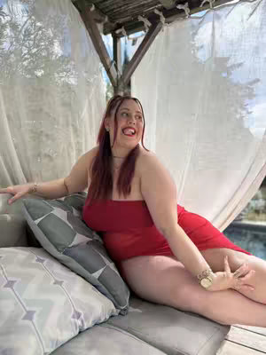 A woman in a red dress reclines on a gray sofa, smiling with one arm resting on a pillow and the other extended. The background includes a wood structure and white curtains.