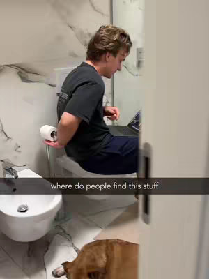 A man sits on a toilet with his laptop, toilet paper roll in hand. Part of a dog is visible in the foreground.