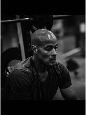 A black and white image of a bald man in a gym setting. The man is looking down at his chest, appearing thoughtful or determined. In the background, gym equipment like a weight rack is visible, though out of focus. The image is composed in shades of grey.