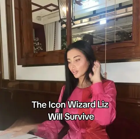 A woman with long dark hair is seated at a table. She is wearing a pink dress, with a two-tone effect. The background shows a mirror and a window. Text overlayed with black font saying 'The Icon Wizard Liz Will Survive'.