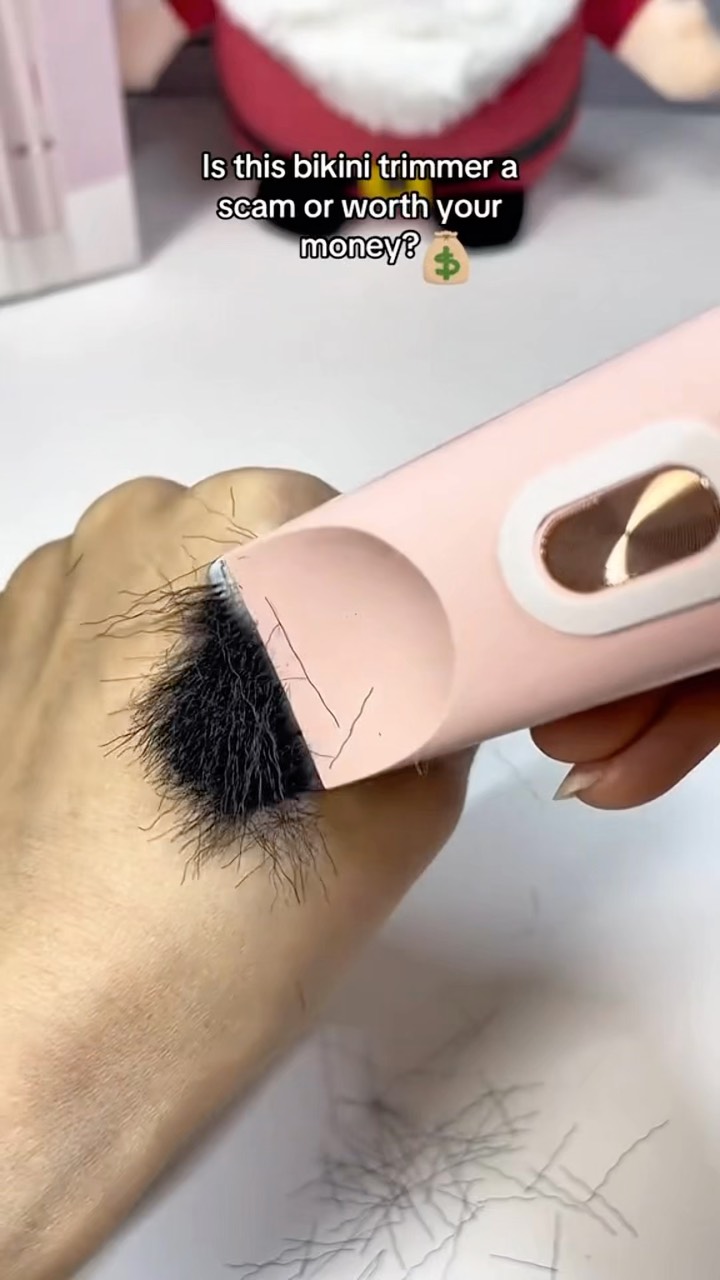 The cover image displays a close-up of a hand with a patch of black hair. A pink electric trimmer is actively cutting the hair, evident from the cut hair falling off. Text layered over the image asks if the bikini trimmer is a scam or worth the money.